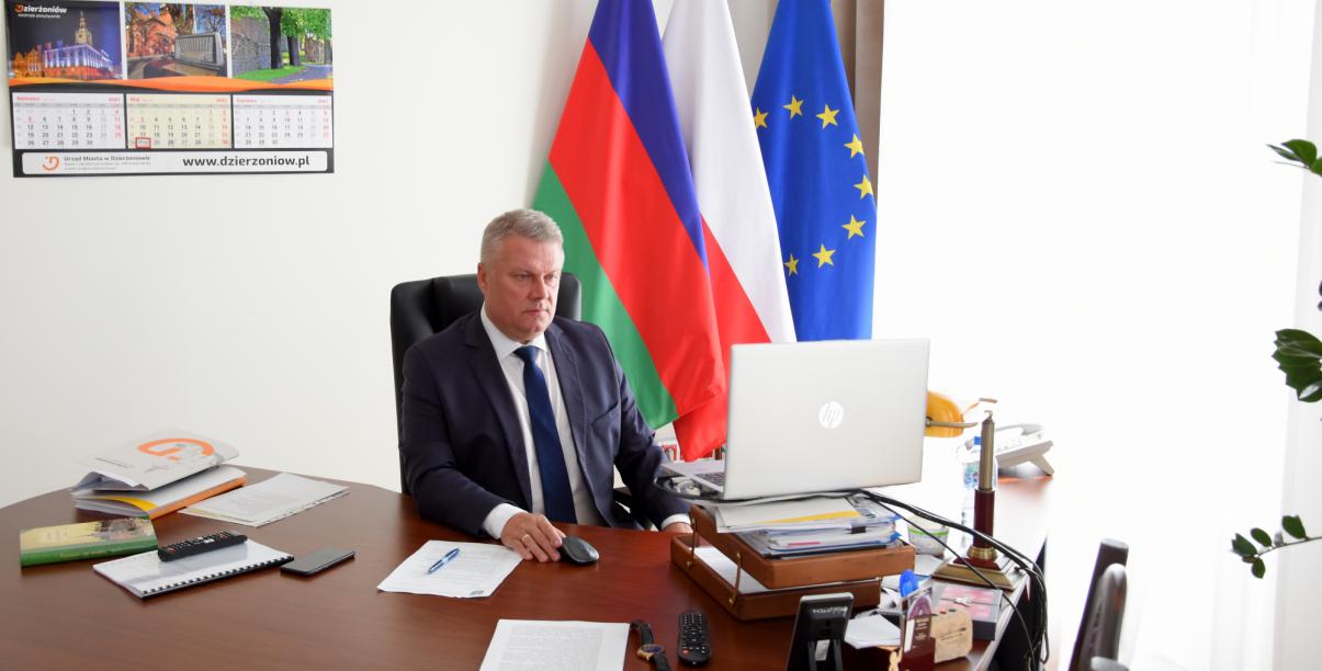 Burmistrz siedzący przy biurku, w tle flagi Polski, UE i Dzierżoniowa