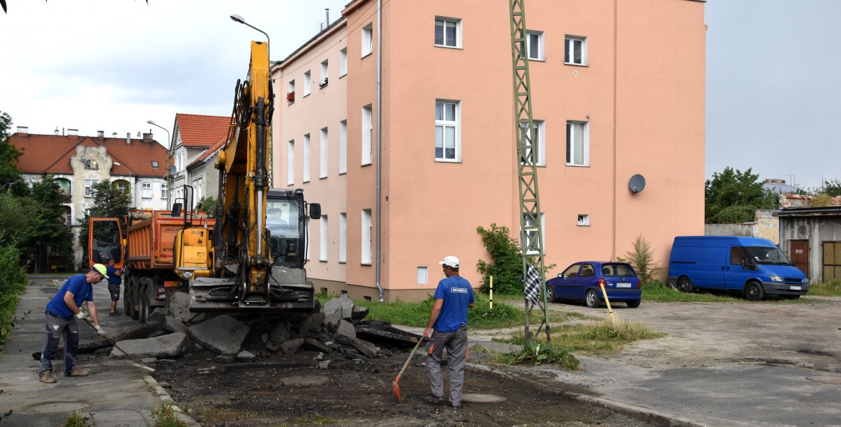 Koparka pracująća na remontowanej drodze, przy koparce pracownik, w tle budynek