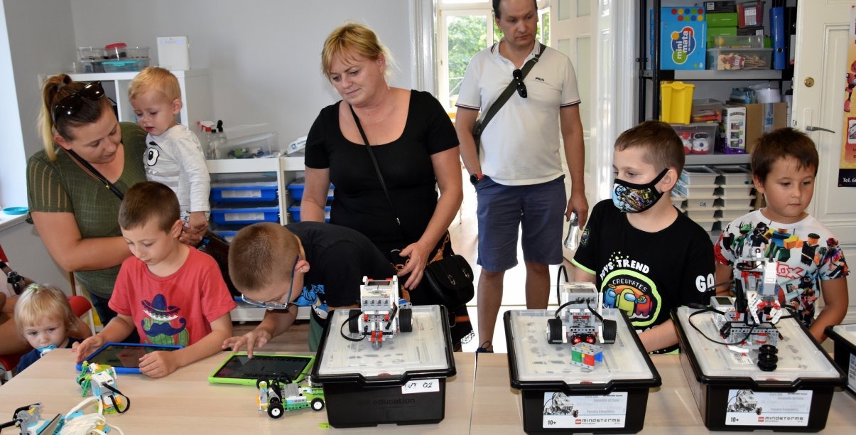 Grupa dzieci i dorosłych przy stołach, na których sa miniaturowe roboty