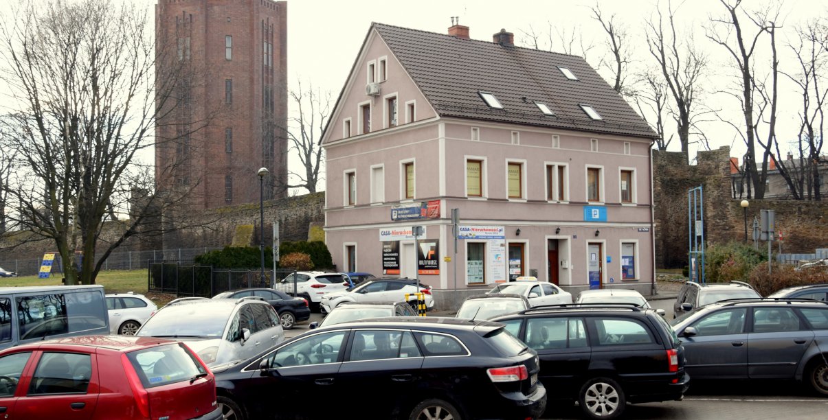 Zaparkowane samochody, w drugim planie kamienica i dalej wieża ciśnień