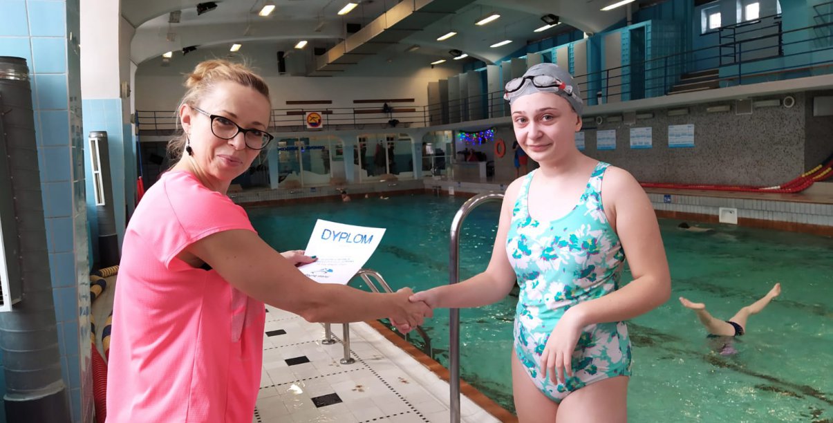 Na zdjęciu intruktor pływania wręcza dyplom dziewczynce, podają sobie dłonie, stoją przy basenie