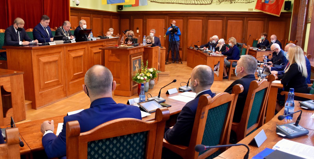 Duża sala, radni siedzący tyłem do obiektywu, w druim planie widok prezydium rady