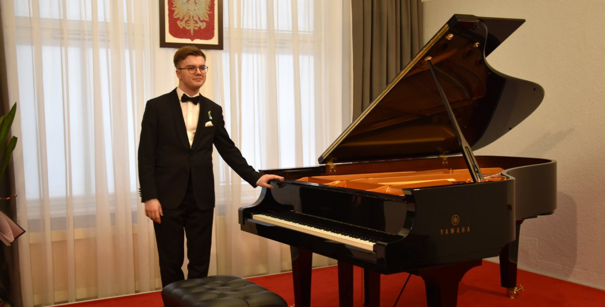 Młody pianista stojący przy fortepianie, z tył godło Polski