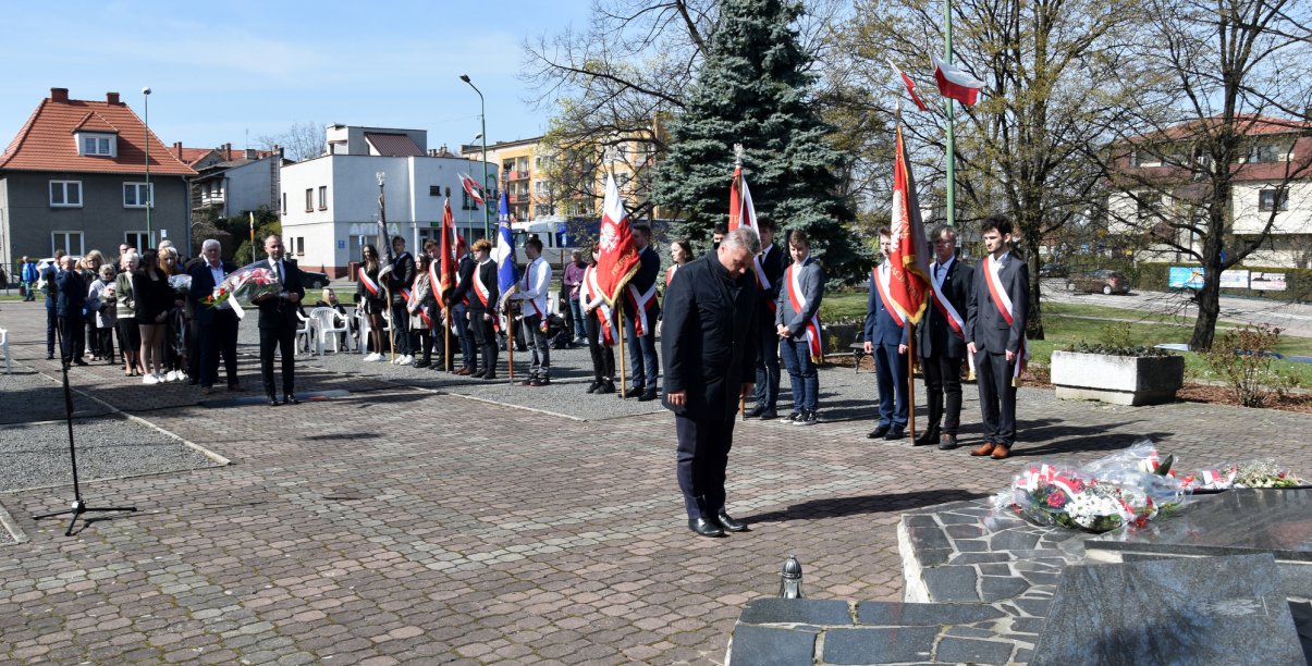 Mężczyzna stojący przed pomnikiem, w durim tle poczet sztandarowy