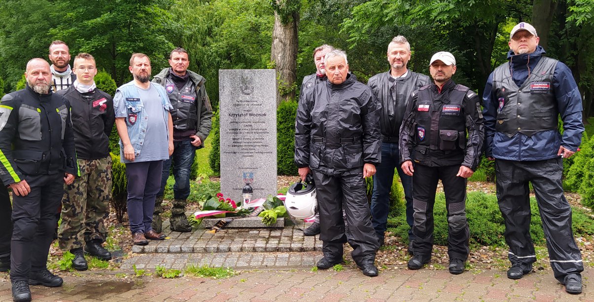 Grupa mężczyzn w strojach motocyklowych stojąca przy kamiennym obelisku 