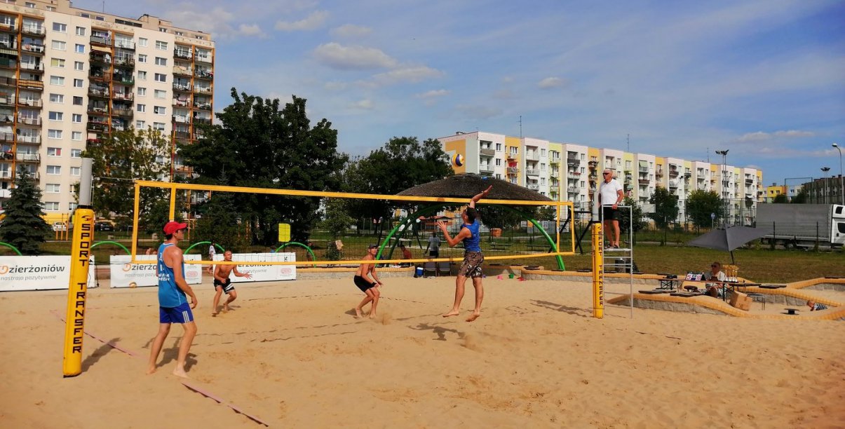 Cztery osoby grają w siatkówke plażową, w drugim tle osiedle mieszkaniowe