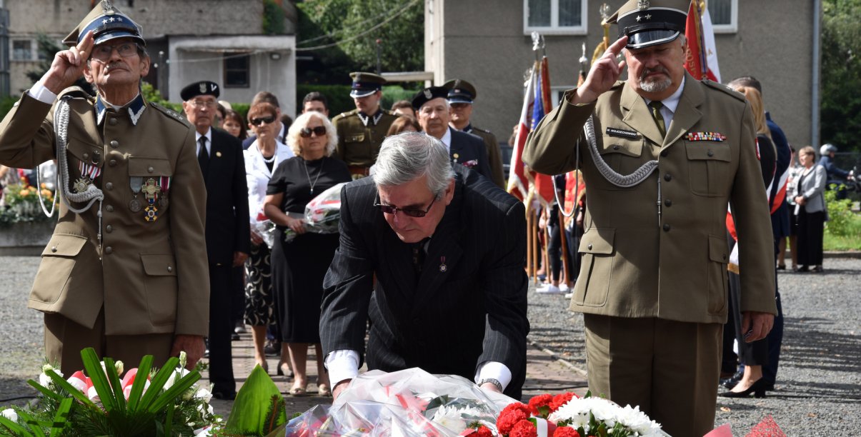 Kombatanci salutujący przy pomniku, na pierwszym planie wieńce kwiatów
