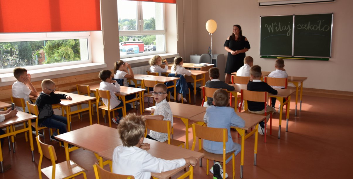 Szkolna klasa, uczniowie siedzący przy łąwkach i uśmiechnięty nauczyciel