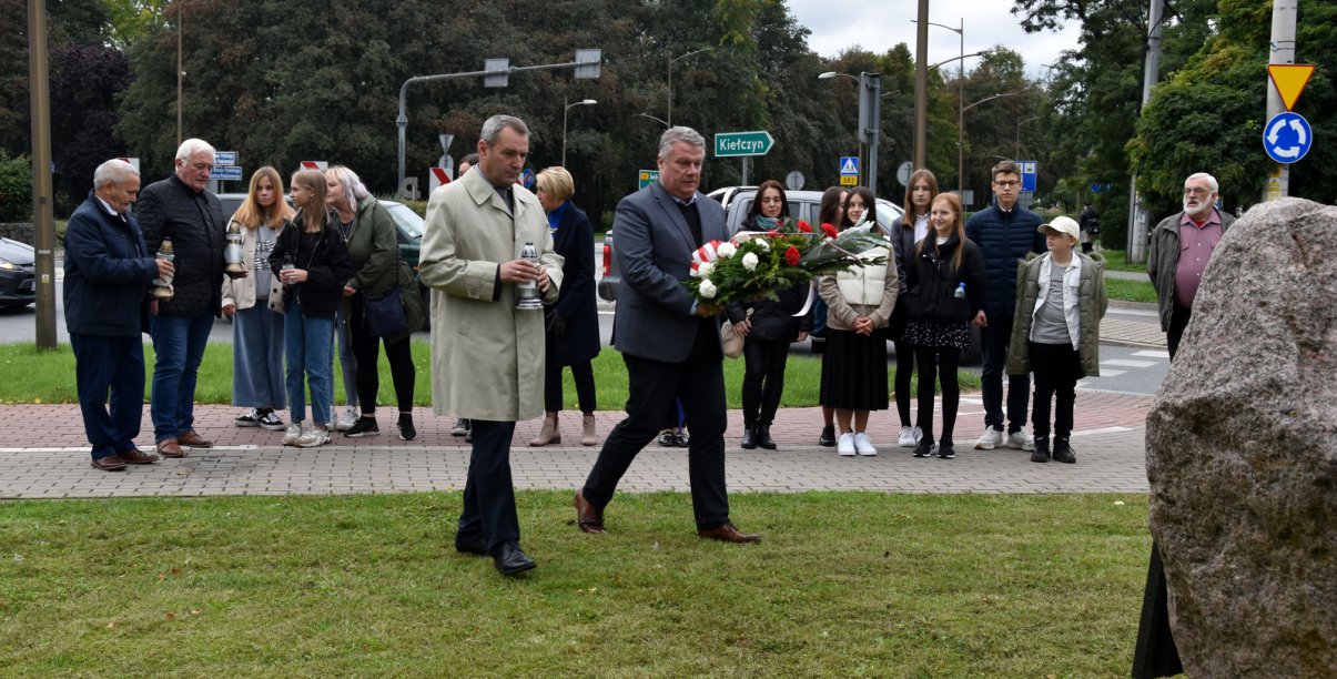 Grupa stojących osób, dwóch panów ze zniczem i kwiatami idą w kierunku kamiennego obelisku