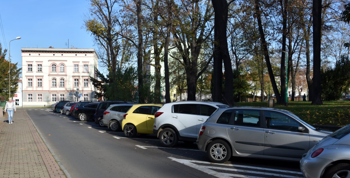 Ulica w mieście, po prawej zaparkowane auta, w drugim planie duża kamienica