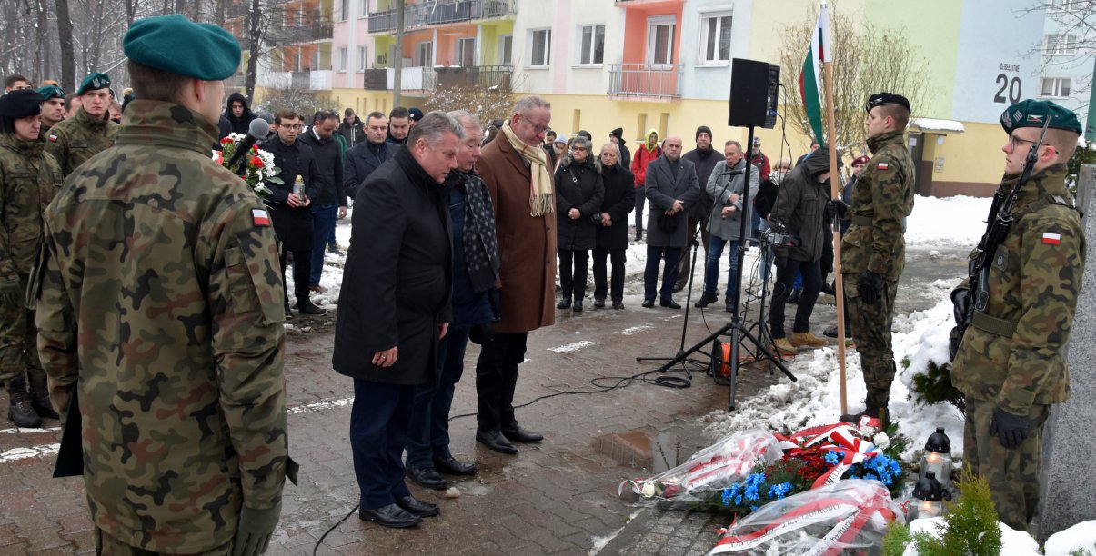 Liczna grupa osób, wśród nich żołnieże, trzy osoby składają ukłon przed obeliskiem