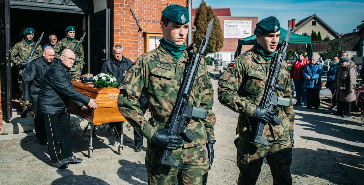 Na pierwszym planie żołnieże, na drugmi grupa osób wynosząca trumnę z kaplicy