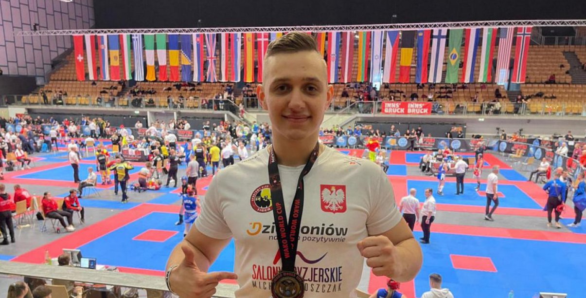 Młody, uśmiechnięty sportowiec, w tle wnętrze hali sportowej z matami do walk