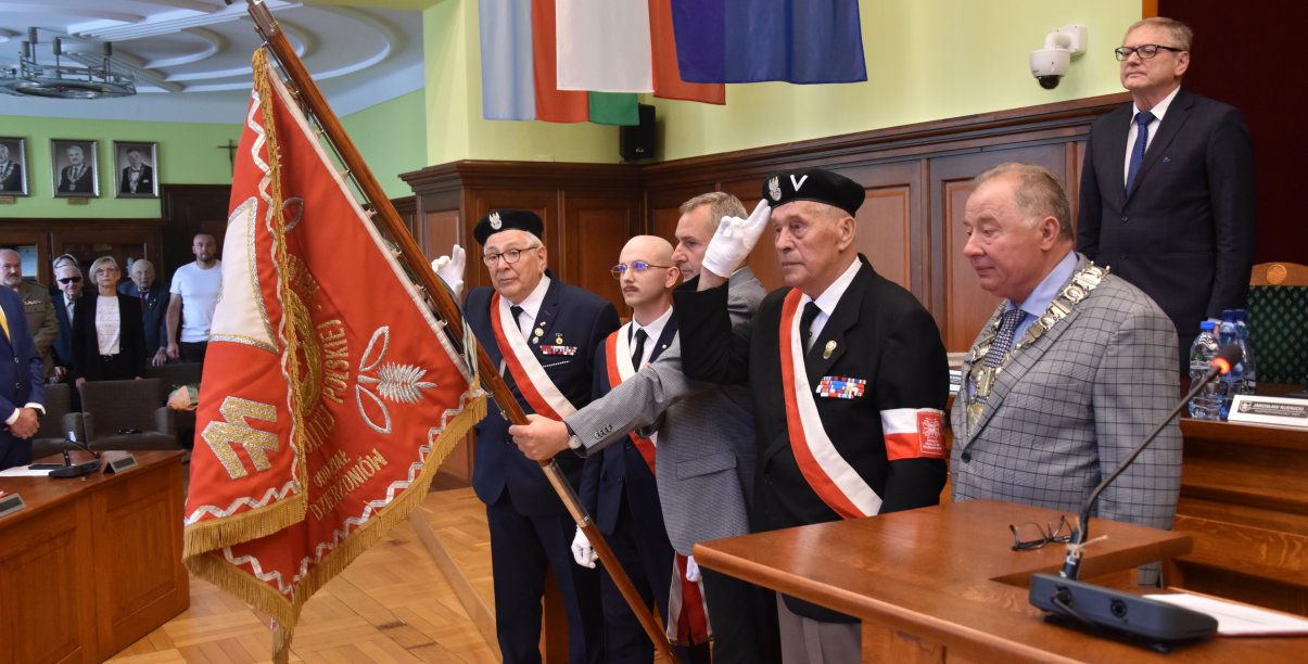 Kombatanci w mundurach trzymają czerwony sztandar i salutują