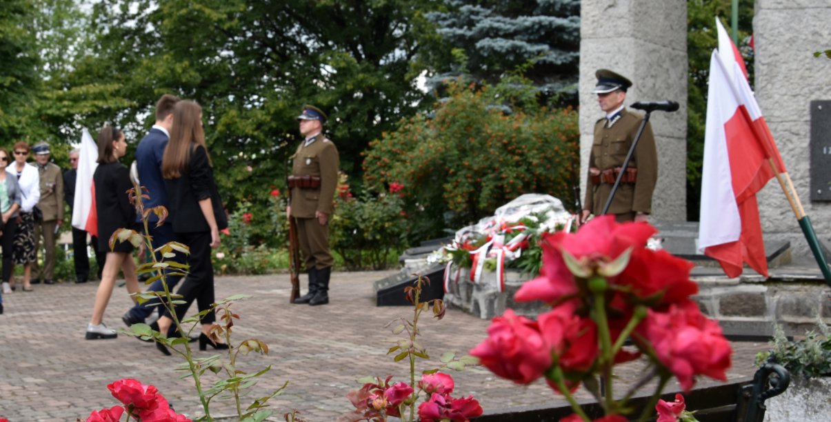 Dwóch żołnieży pełni wartę przy pomniku, do któego podchodzi grupa młodzieży z kwiatami, obok flaga Polski
