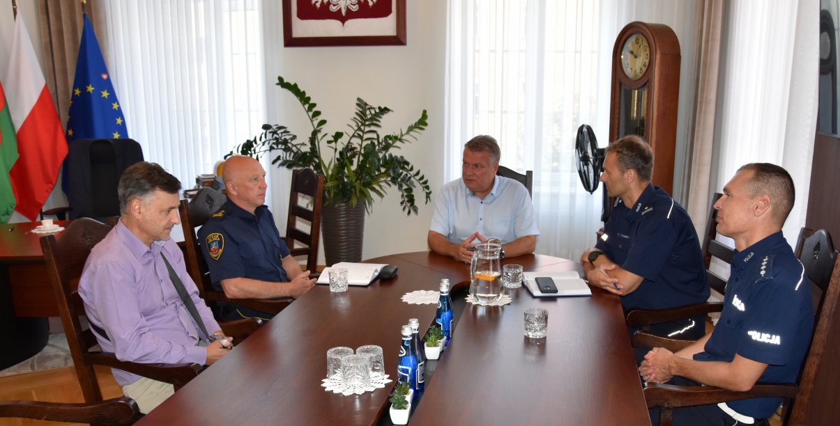 Pięciu mężczyzn przy dużym stole, po prawej dwóch policjatów, po lewej straznik miejski i osoba po cywilnemu, u szytu stołu mężczyzna w białej koszuli