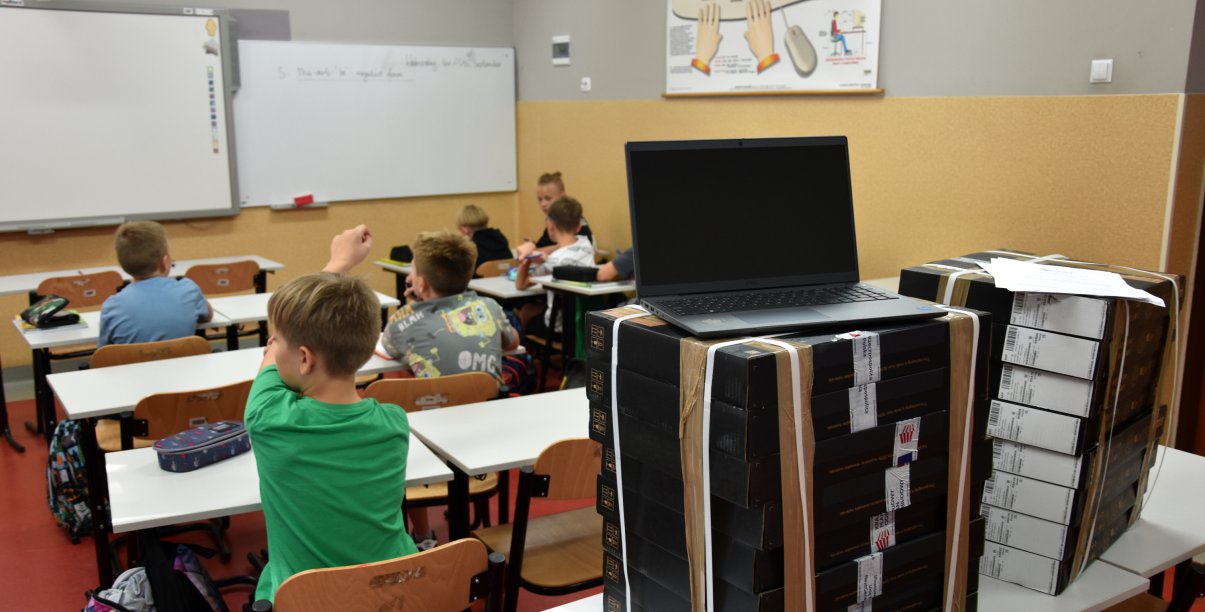 Uczniowie podczas zajęć w klasie, na pierwszym planie laptop i uczeń z podniesioną ręką 