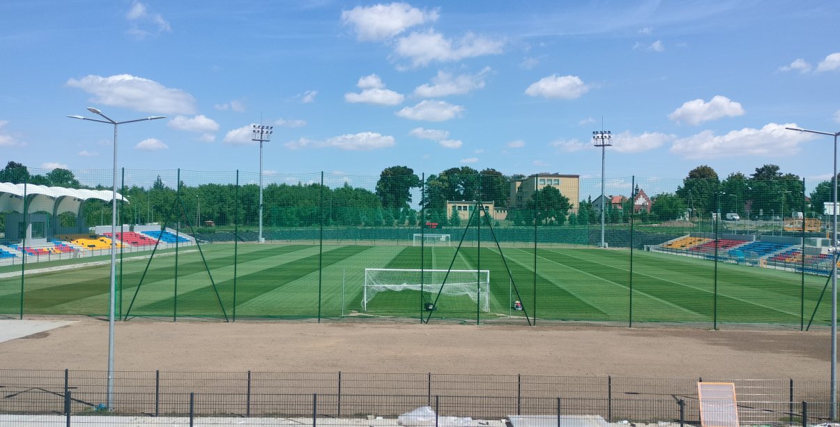 Stadio, widok z góry od strony bramki. Po bokach kolorowe trybuny, w tle zieleń drzew