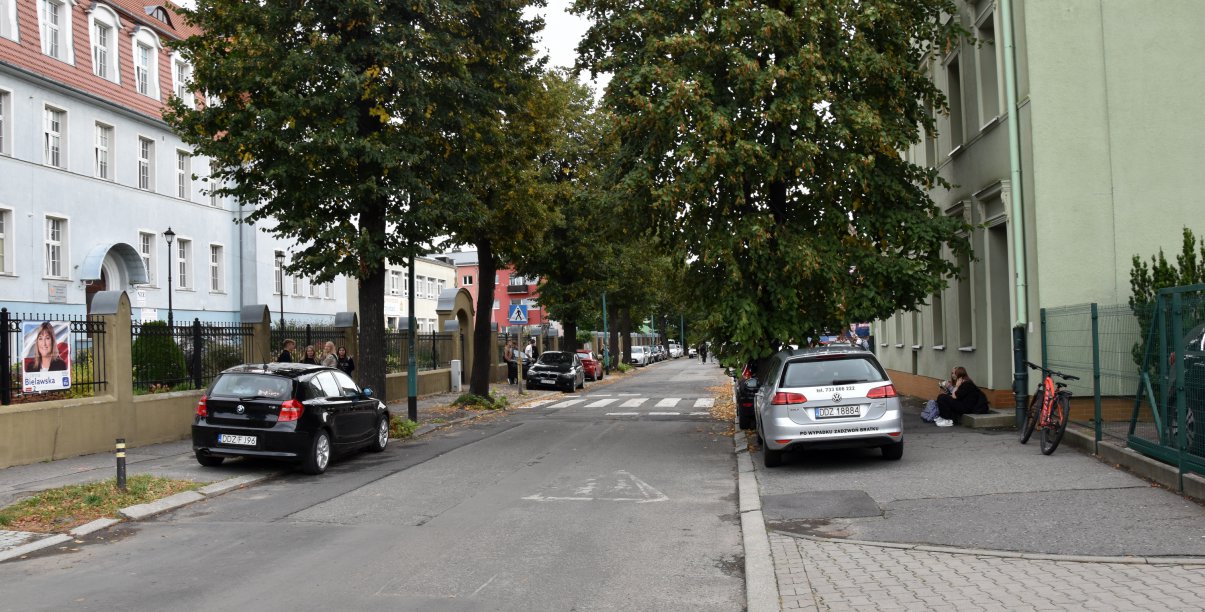 Ulica, zaprakowane na chodniach auta, w tle kamienice i zieleń 