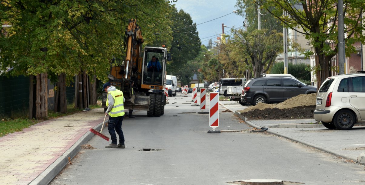 Prace przy budowie drogi, człowiek w jaskrawej kamizelce zamiata drogę, za nim koparka