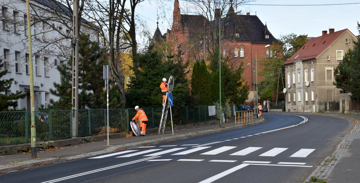 Droga, przejśćie dla pieszych, dwie osoby w pomarańczowych strojach montują znak drogowy