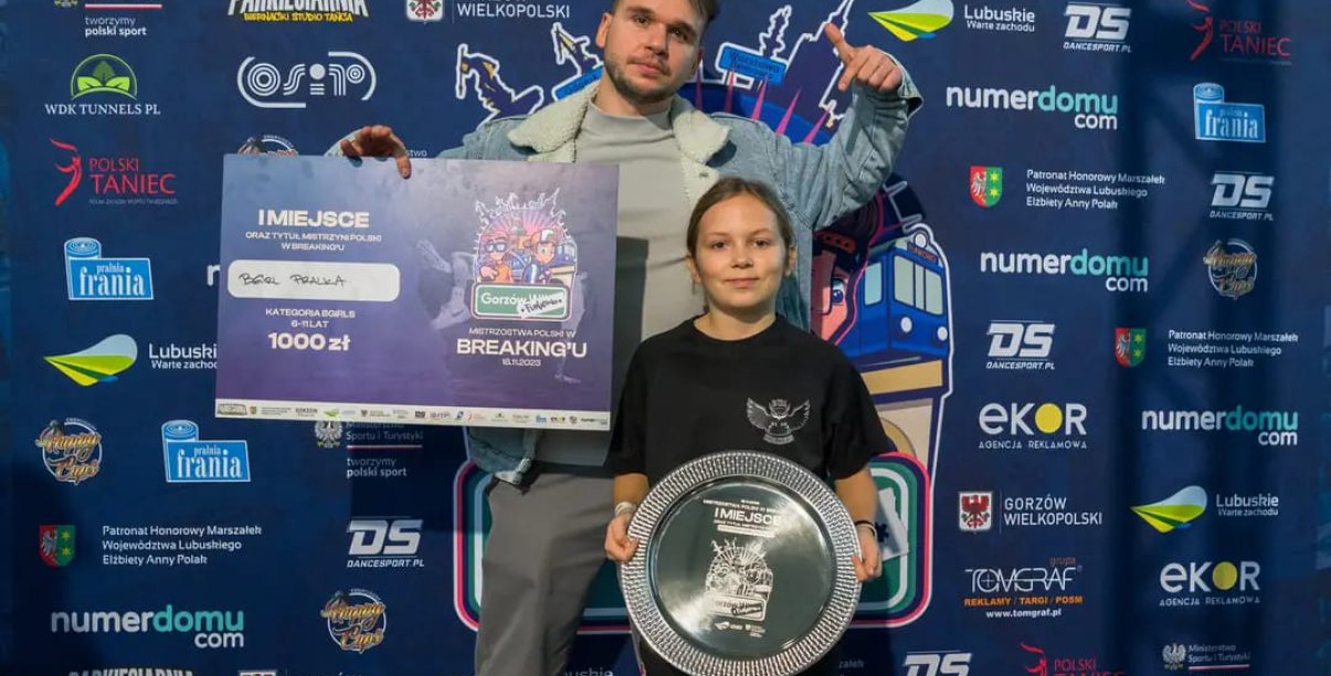 Trener trzyma w reku duży czek, przed nim zawodniczka ze srebrną paterą w tle ścianka promocyjna