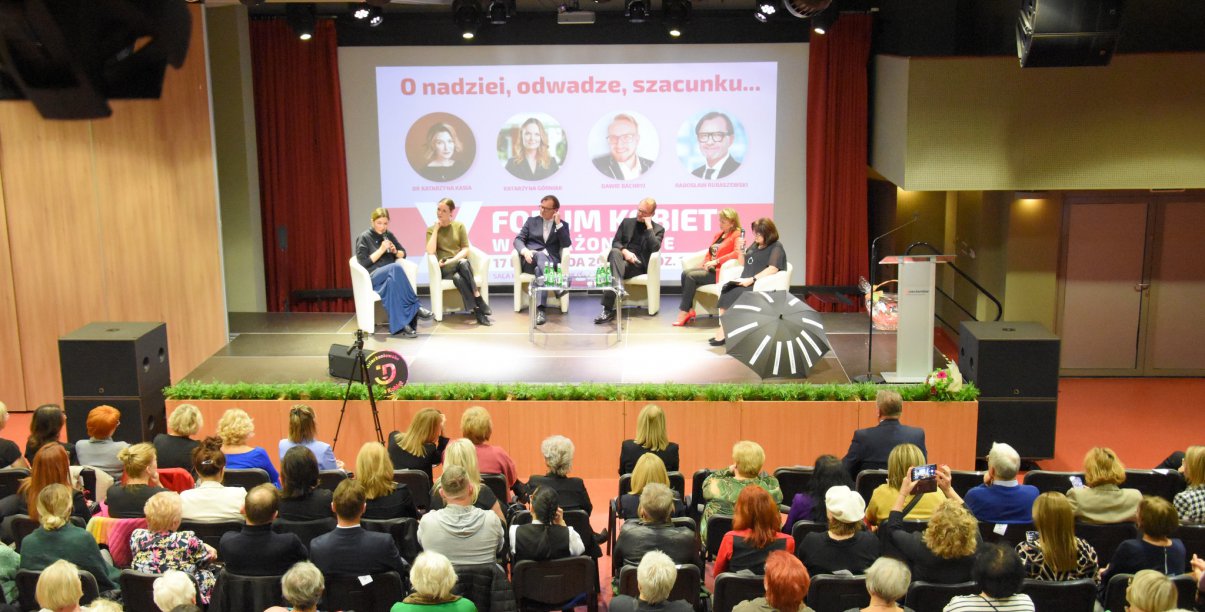 Sala, na scenie osoby siedzące w fotelach, przed nimi publiczność