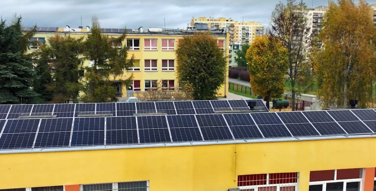 Panele fotowoltaiczne na dachu budynku, w tle osiedle mieszkaniowe