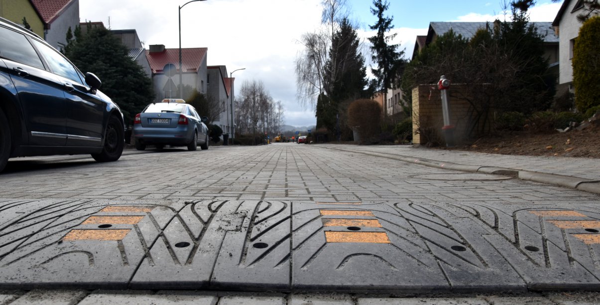 Droga z kostki betonowej i widoczny na niej "spowalniacz", o lewej dwa zaparkowane auta