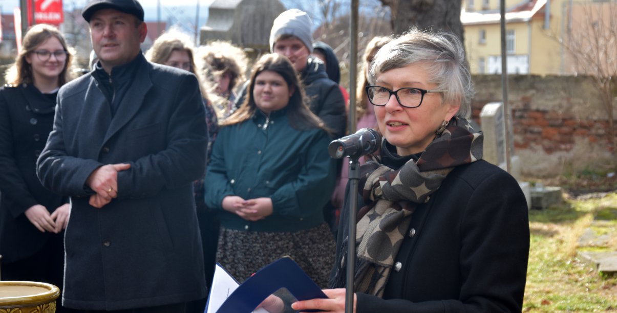 Kobiet mówiąca do mikrofonu, za nią grupka ludzi