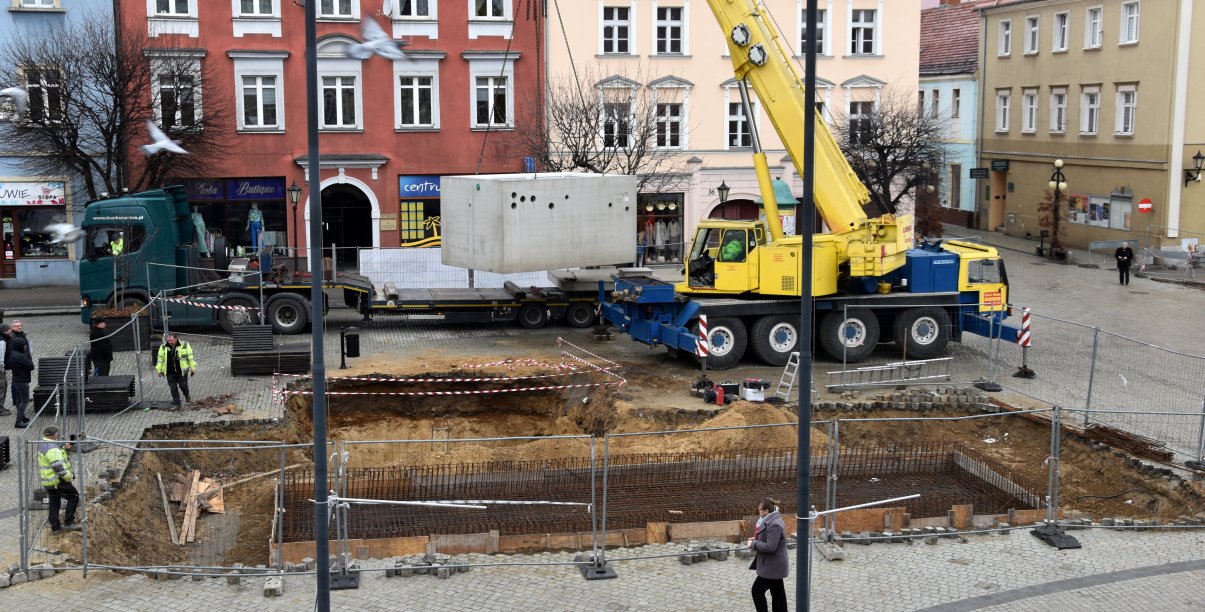 Dźwig unoszący dużą betonową konstrukcję, w tle kamienice