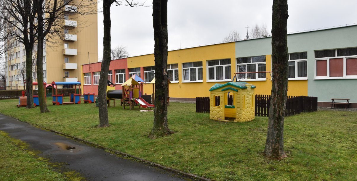 Kolorowy, podłużny parterowy budynek żlobka, przed nim plac zabaw i teren zielony