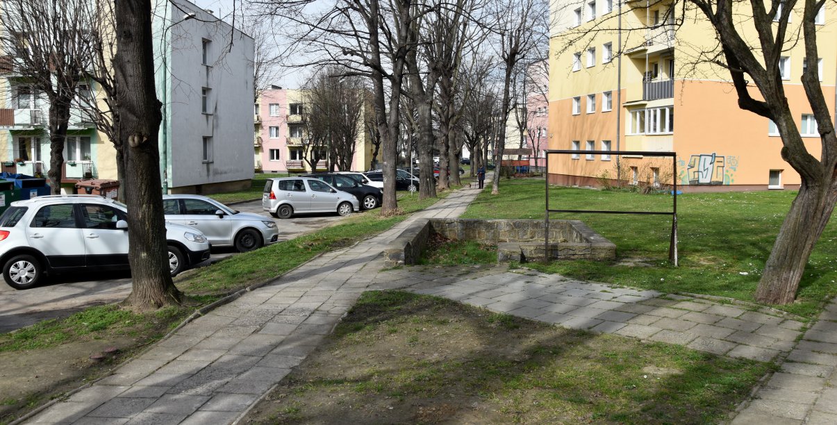 Osiedle, chodnik, obok zaparkowane auta,. w drugim planie bloki mieszkalne