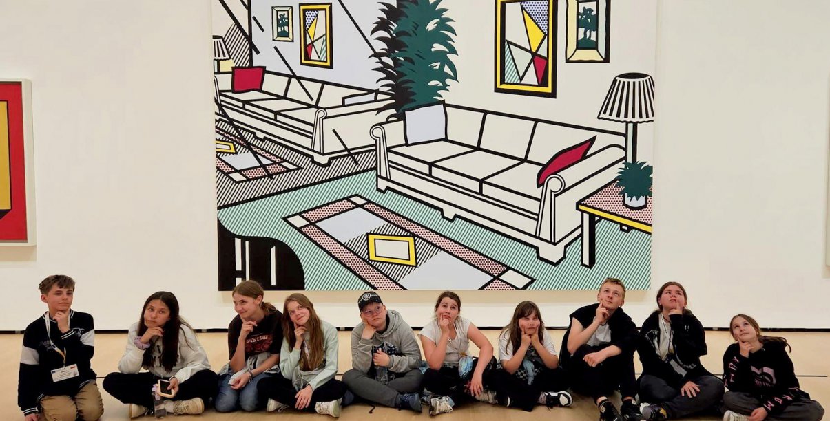 Dzisięcioro uczniów siedzi na parkiecie, za nimi duży obraz przedsawiającyc pokój 