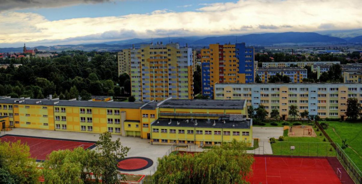 Budynek szkoły z góry, przed nim dwa czerwone boiska sportowe, za budynkiem bloki i wieżowce oseidlowe, w dalekim planie góry