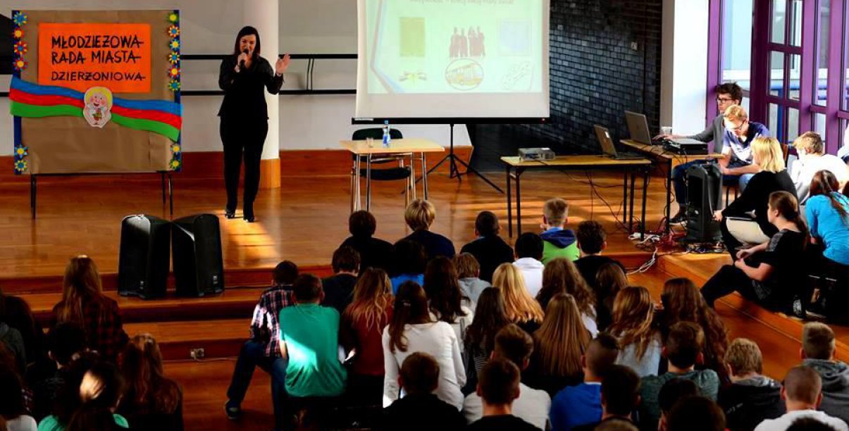 Aula szkolna, dziewczyna stoi przed dużą grupą uczniów i mówi do nich przez mikrofon