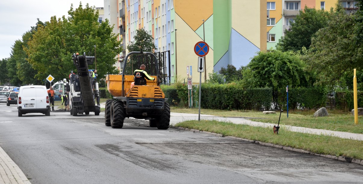 Droga osiedlowa, na części jezdni maszyny do prac drogowych w tle kolorowy blok