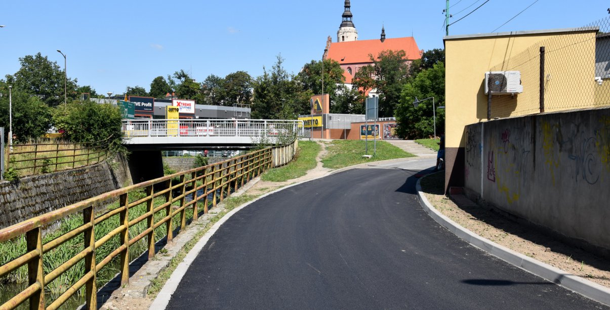 Droga z nowym asfaltem, po lewej kamienne koryto rzeki, po prawej mur, w drugim planie średniowieczny kościół