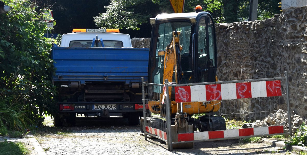 Mała koparka i mały samochód dostawczy na wąskiej drodze, obok drogi kamienny mur