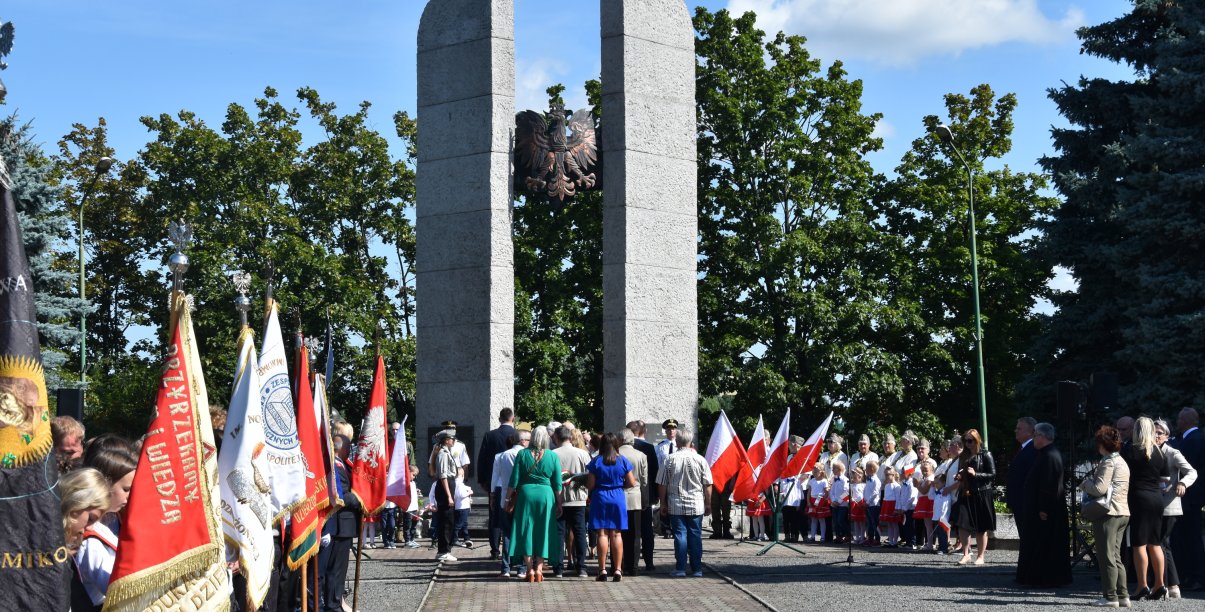 Grupa ludzi stojąca przed pomnikiem, obok poczet sztandarowy i młodzież w białych koszulach 