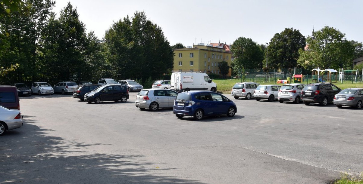 Asfaltowy parking z zaparkowanymi autami, po prawej stronie budynki