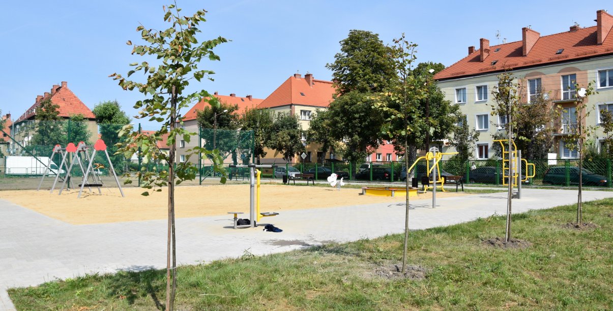 Plac zabaw z dużą piaskownicą, na pierwszym planie drzewka, w tle budynki mieszkalne