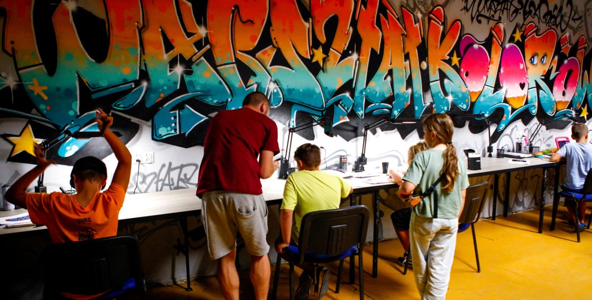 Na tle kolorowej ściany graffiti dzieci siedzą przy stołach i rysują, obok stoi instruktor