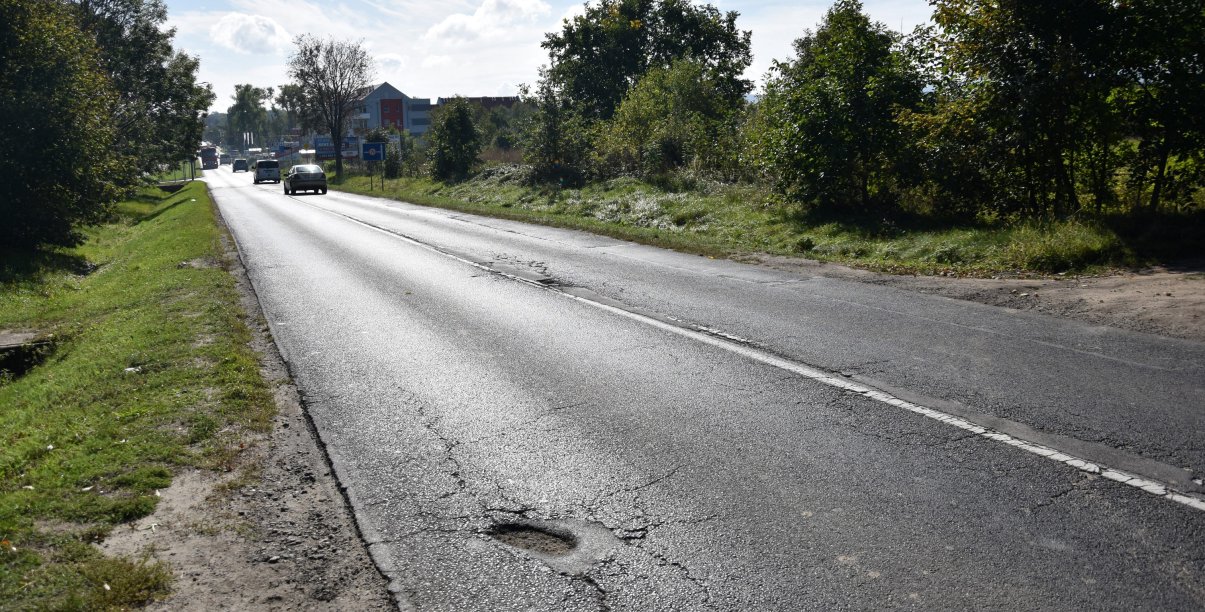 Droga asfaltowa prowadząca do miasta, w dali samochody, po bkach drzewa i krzewy