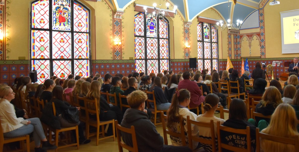 Aula szkolna z dużymi okiennymi witrażami zapełniona uczniami