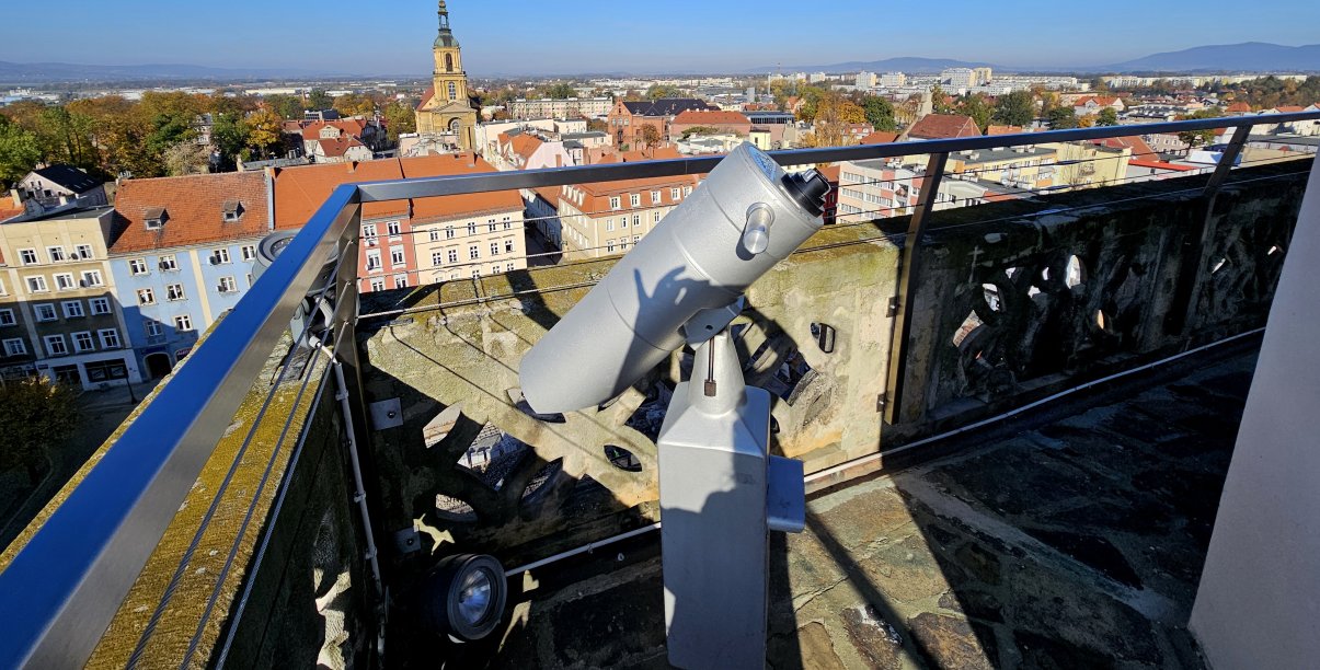 Wieża ratuszowa z kaminnymi i metalowymi barierkami, na pierwszym planie luneta, w tle panorama miasta