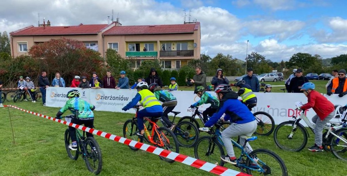 Siedmiu chłopaków ścigających się na rowerach po wyznaczonej trasie, na drugim planie zieleń i budynki