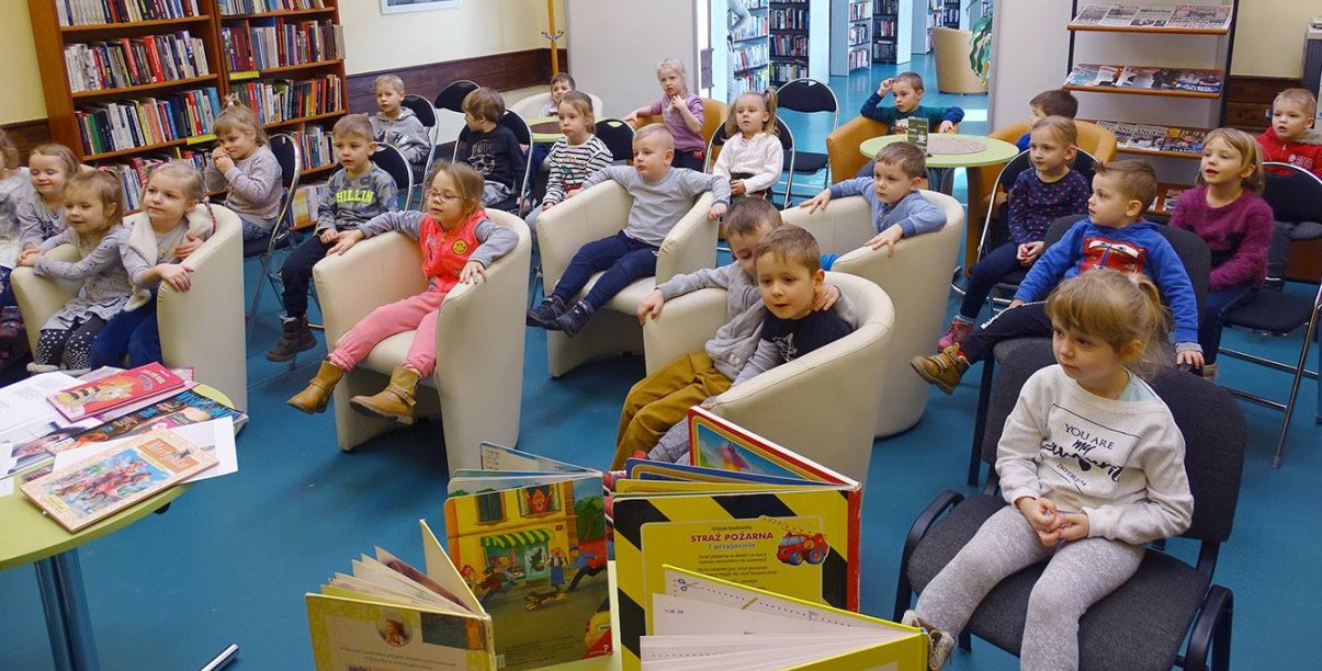 Przedszkolaki siedzące w fotelach w pomieszczeniu biblioteki, na pierwszym planie książki