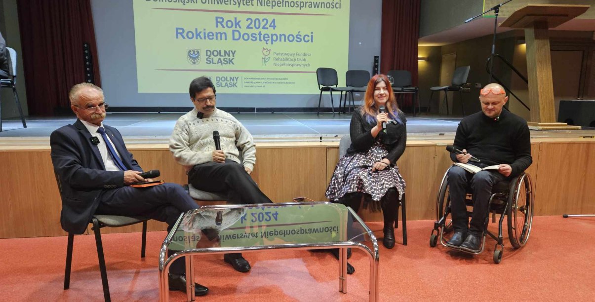 Trzy osoby siedzące na krzsłach, jedna na wózku inwalidzkim, przed nimi szklany stolik