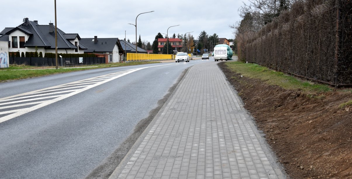 Droga i chodnik z kostki betonowej, po prawej stronie żywopłot, po lewej dom jednorodzinny